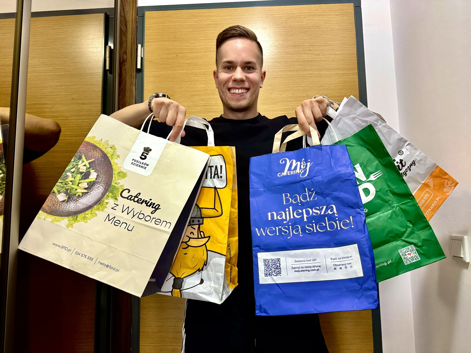 Adrian Domagalski, dietetyk z Poznania, trzyma torby z dostawami cateringów z testu: 5 Posiłków Dziennie, Kuchnia Vikinga, Mój Catering, Robin Food i Tytka Fit