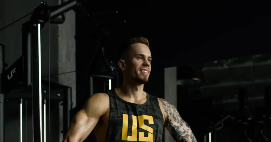 Adrian Domagalski w tank topie Make Us Strong w kolorze moro z żółtym logo US, na siłowni