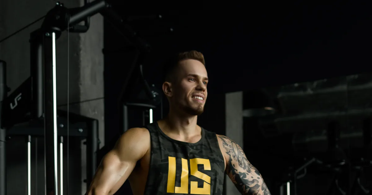 Adrian Domagalski w tank topie Make Us Strong w kolorze moro z żółtym logo US, na siłowni