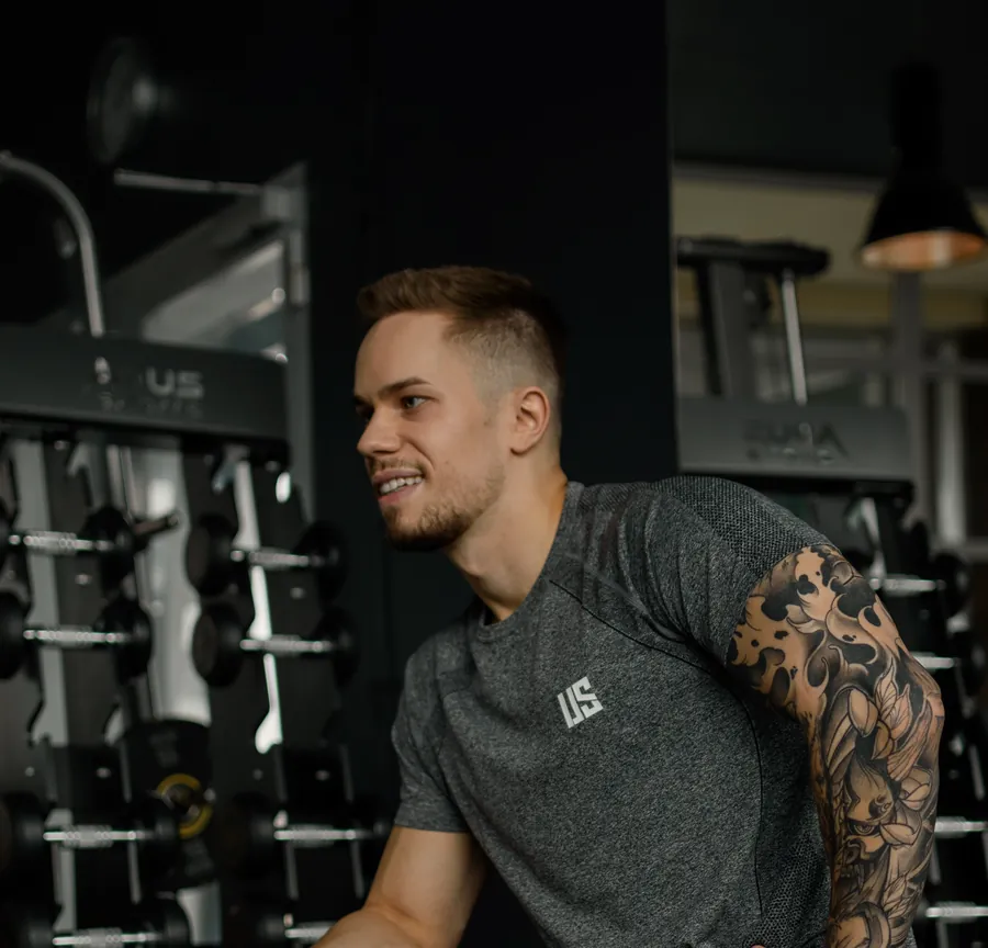 Adrian Domagalski w szarej koszulce Make Us Strong z białym logo US, na siłowni