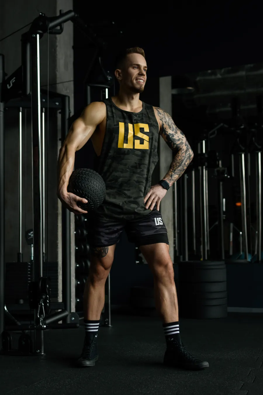 Adrian Domagalski w tank topie Make Us Strong moro z żółtym logo US i czarnych spodenkach z logo US, z piłką slam ball