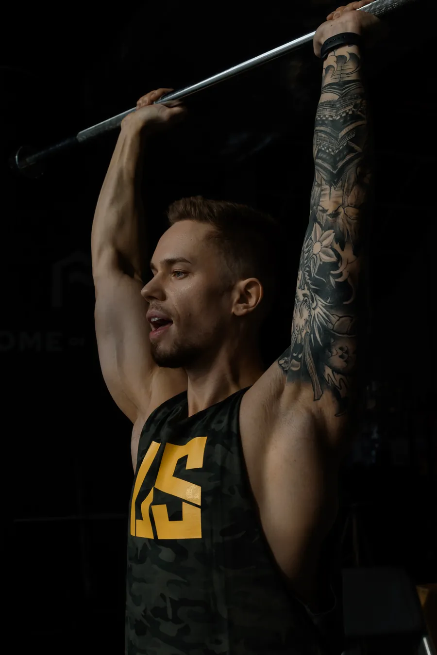 Adrian Domagalski w tank topie Make Us Strong moro z żółtym logo US trzyma gryf nad głową, w tle logo studia Home of Body